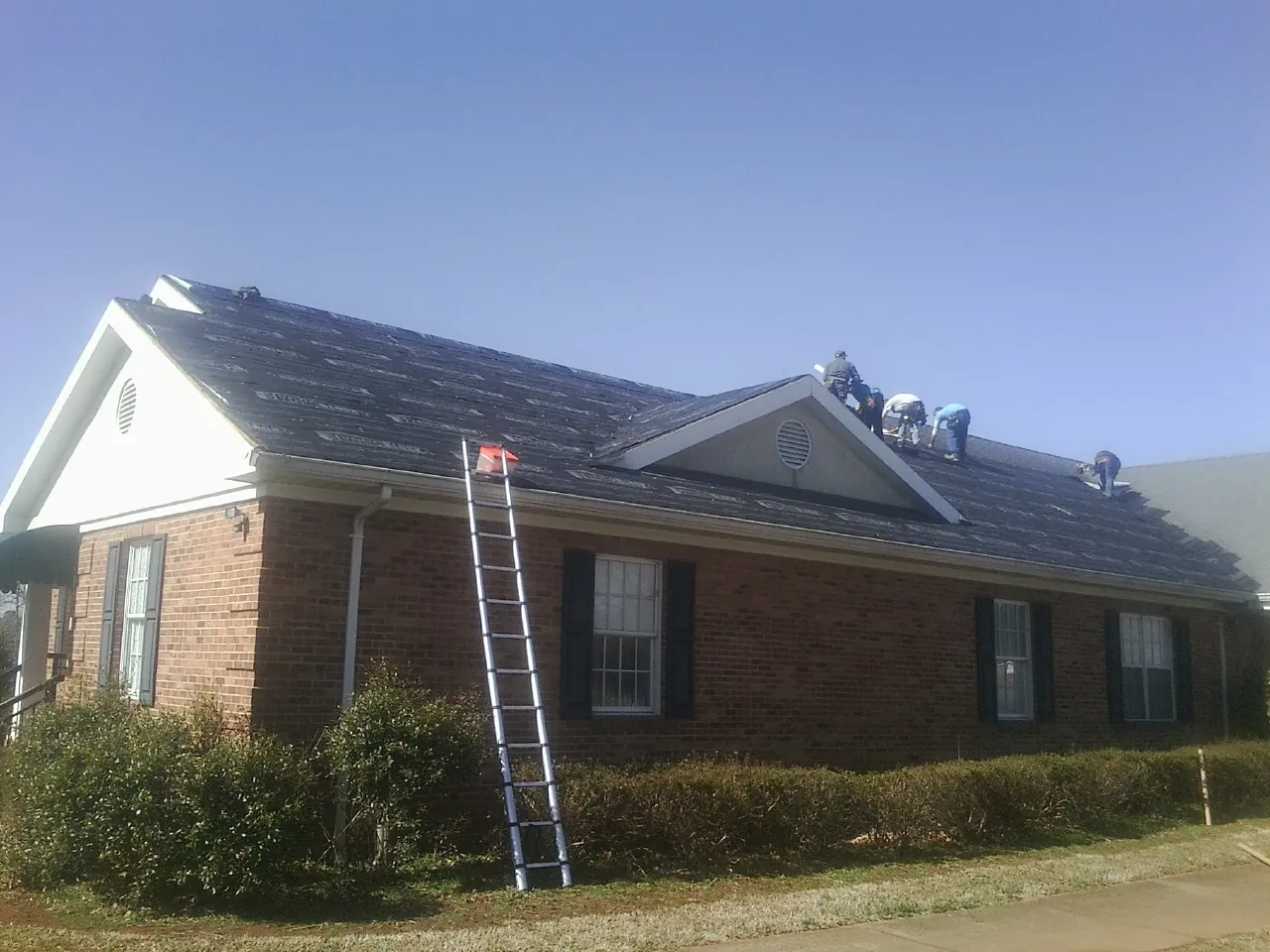 Crew tearing shingles off a brick ranch roof with a ladder set — Emergency Roof Repair work by our crew in Hartly