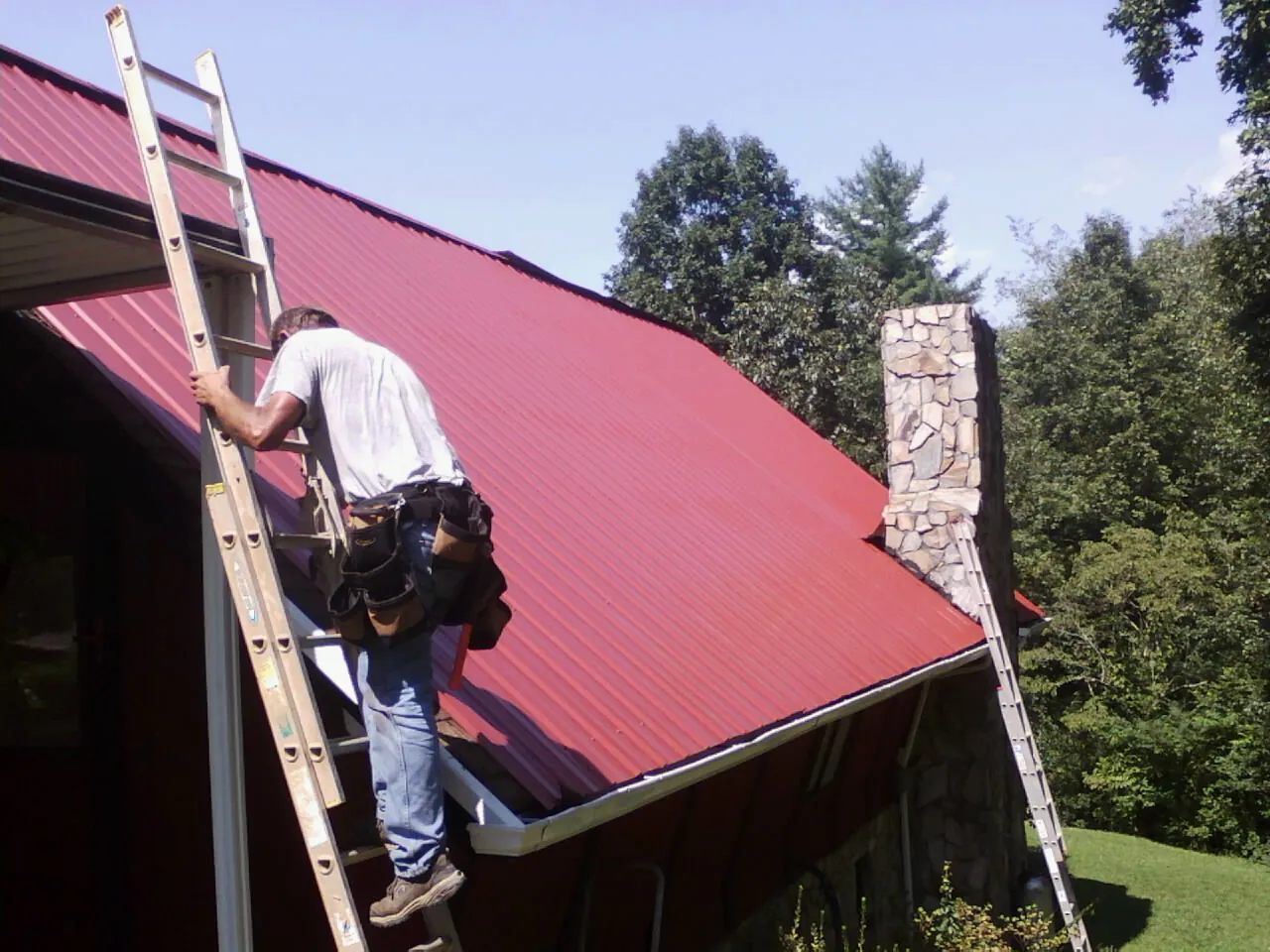 Roofer on a ladder beside a red metal roof and stone chimney — Soffit & Fascia Repair by Roofing Brothers in Hartly